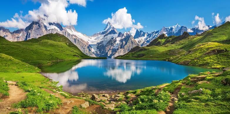 A wide alpine vista — green meadows, mirror lake, snow-capped peaks